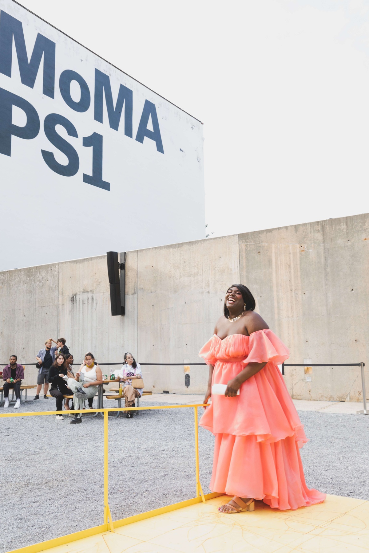 A Black trans woman joyfully walks onto a yellow stage in flowing pink dress. She is in a courtyard with a wall in the background that reads "MoMA PS1".