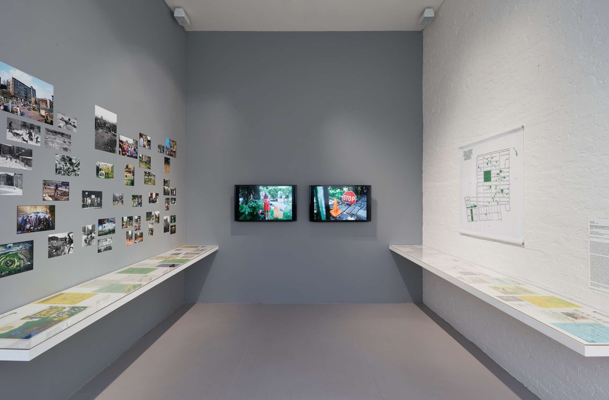 A white and grey walled gallery space with two TV’s on the center wall, vintage images on the left wall, and a floor plan on the right wall. 