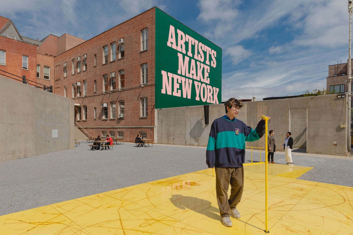 A person in a hip outfit stands on a yellow stage holding a yellow mic. In the distance people sit at picnic tables. A mural behind the people reads "ARTISTS MAKE NEW YORK" in large light pink letters on a forest green background.