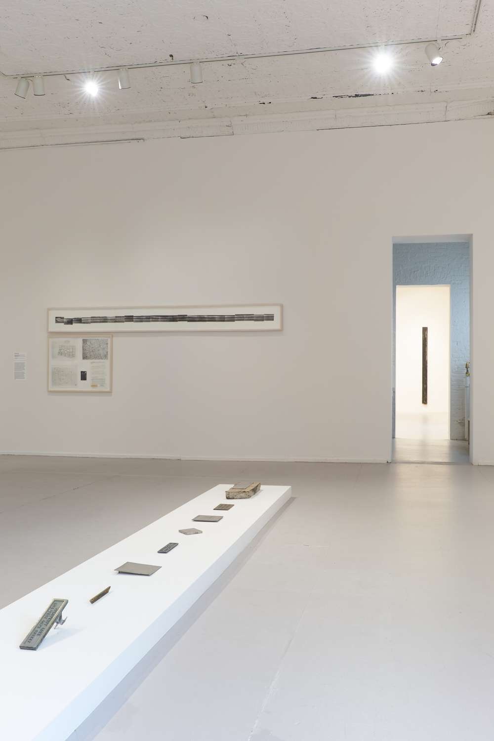 A gallery space with industrial looking objects on top of a long pedestal and long black and white artwork in the background.