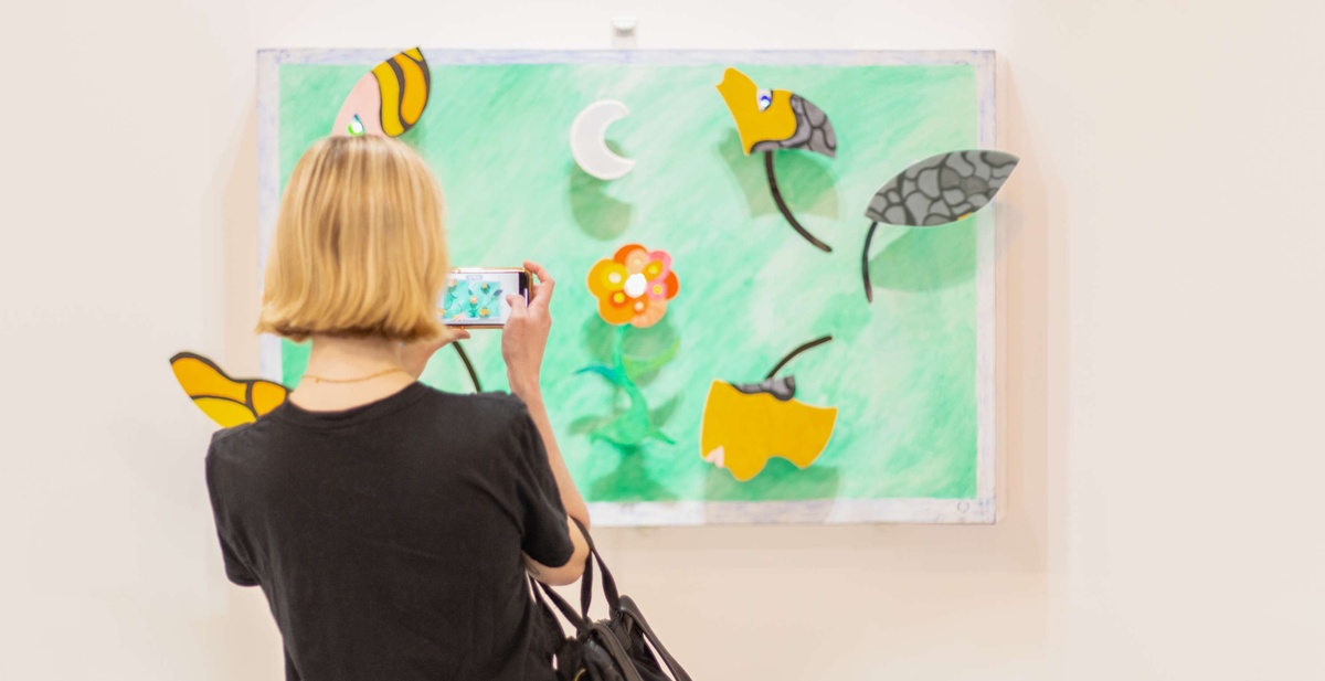 A person uses a phone to take a picture of an artwork with a bright mint green background in which black, yellow, grey, and beige abstract leaf-like shapes pop off the canvas.