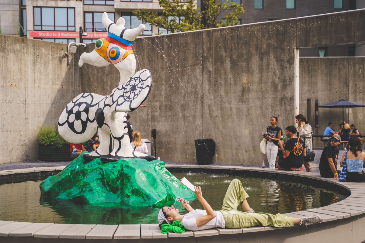 A person reading a book lays on the edge of a circular fountain. There is a sculpture in the center of a fountain with a female figure in a colorful striped leotard spewing water.