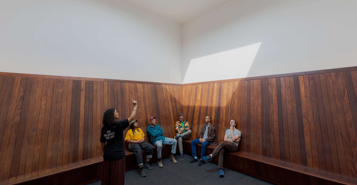 A group of people sit in the corner of a room on a wood-paneled bench. A person points upward as the light from a hole in the ceiling creates a hexagonal shape that falls on the people.
