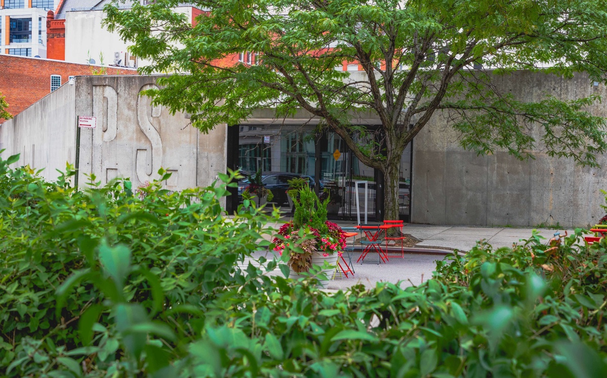 A view outside of the MoMA PS1 entrance with green foliage in the foreground. A pot of plants and red flowers sits near blue and red metal tables and chairs.