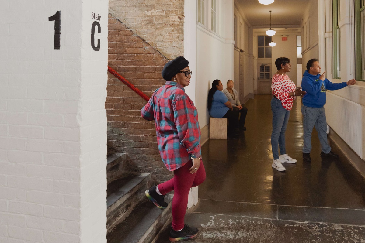 A visitor steps down a stairwell onto the first floor of MoMA PS1, coming into view of other visitors in the hallway who sit on a bench and gesture out the window.