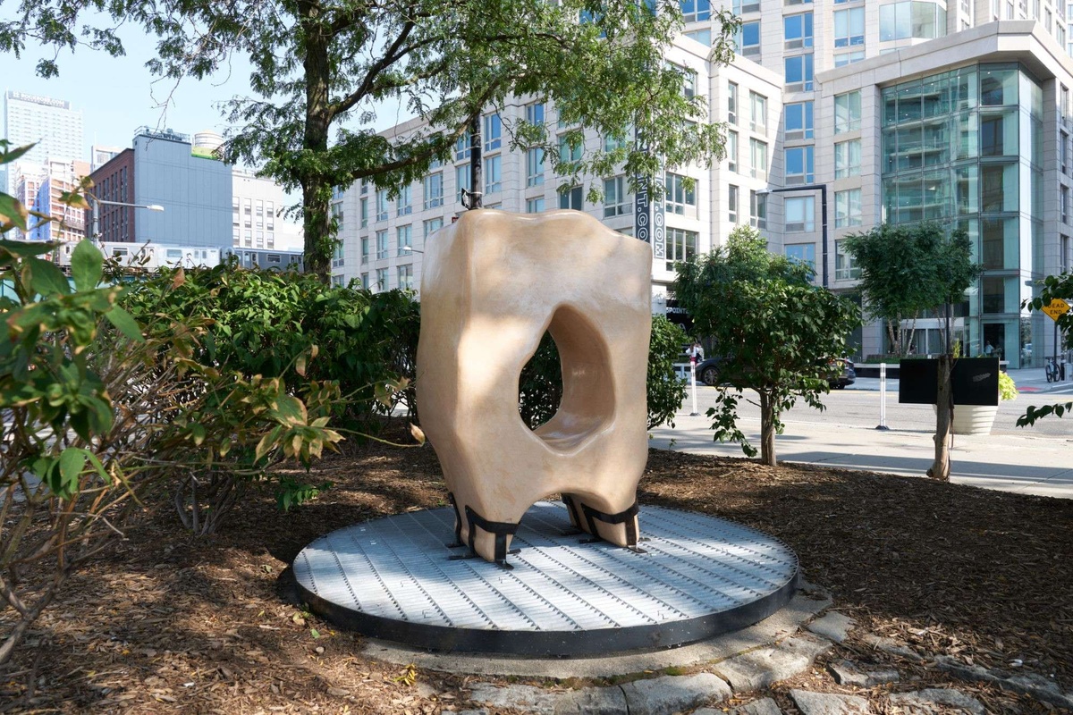 A brown abstract ceramic sculpture, which recalls a misshapen donut, sits in a small park in front of a city street,
