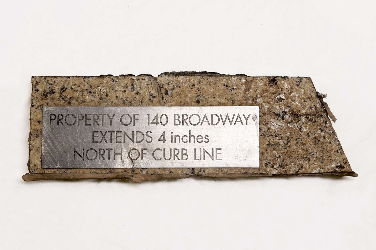 A close up image of a stone textured paper with a silver plaque with the engraving "PROPERTY OF 140 BROADWAY EXTENDS 4 inches NORTH OF CURB LINE."
