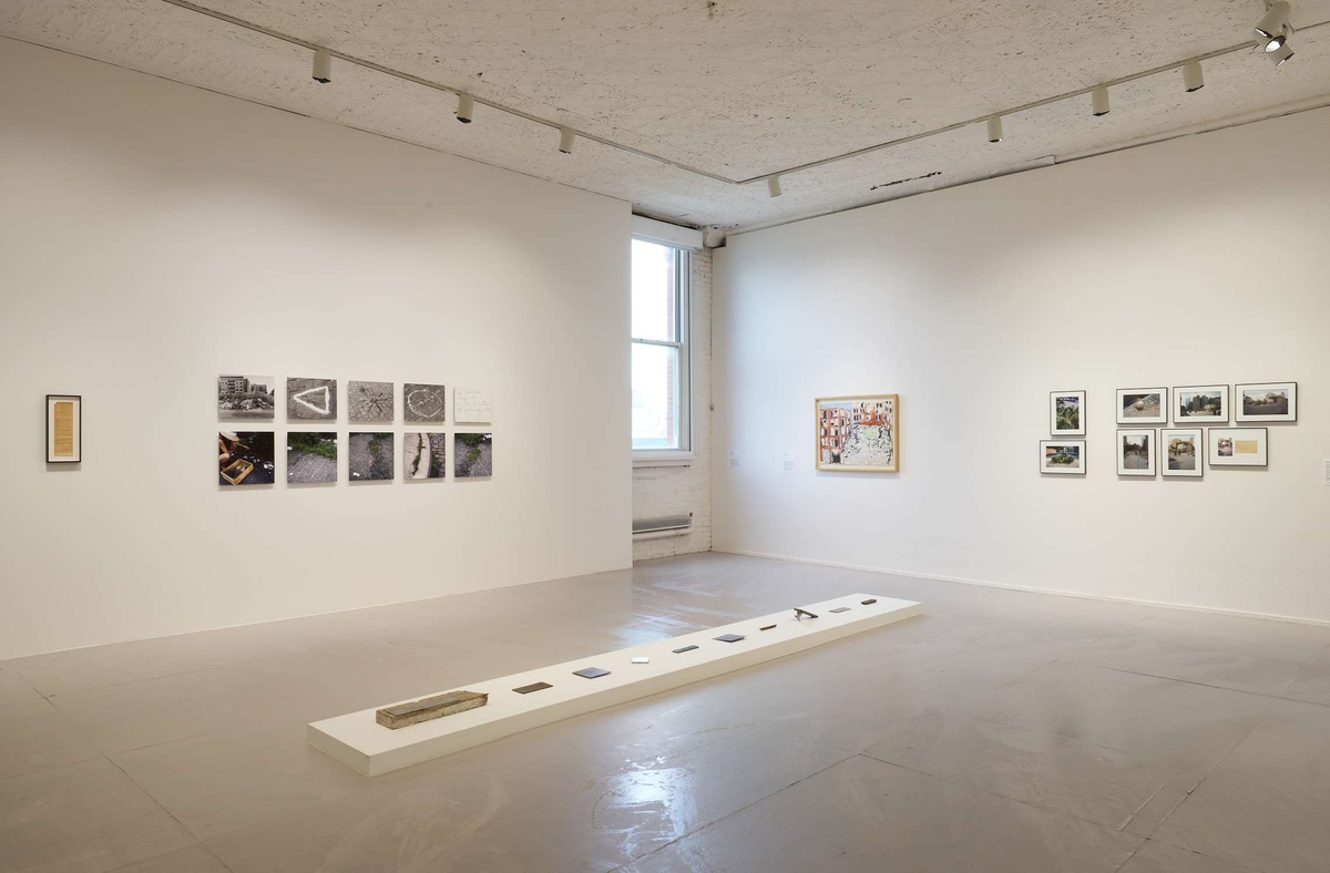 A corner gallery with a long, short, pedestal in the center of the floor with found industrial objects on it. Groups of building artwork adorn the walls behind it.