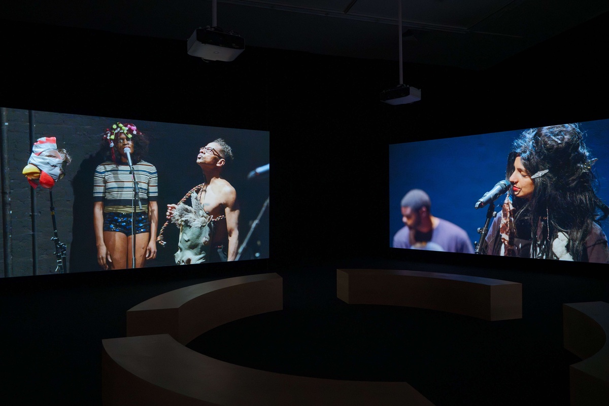 Two screens display pre-recorded dance performances in a dark room.