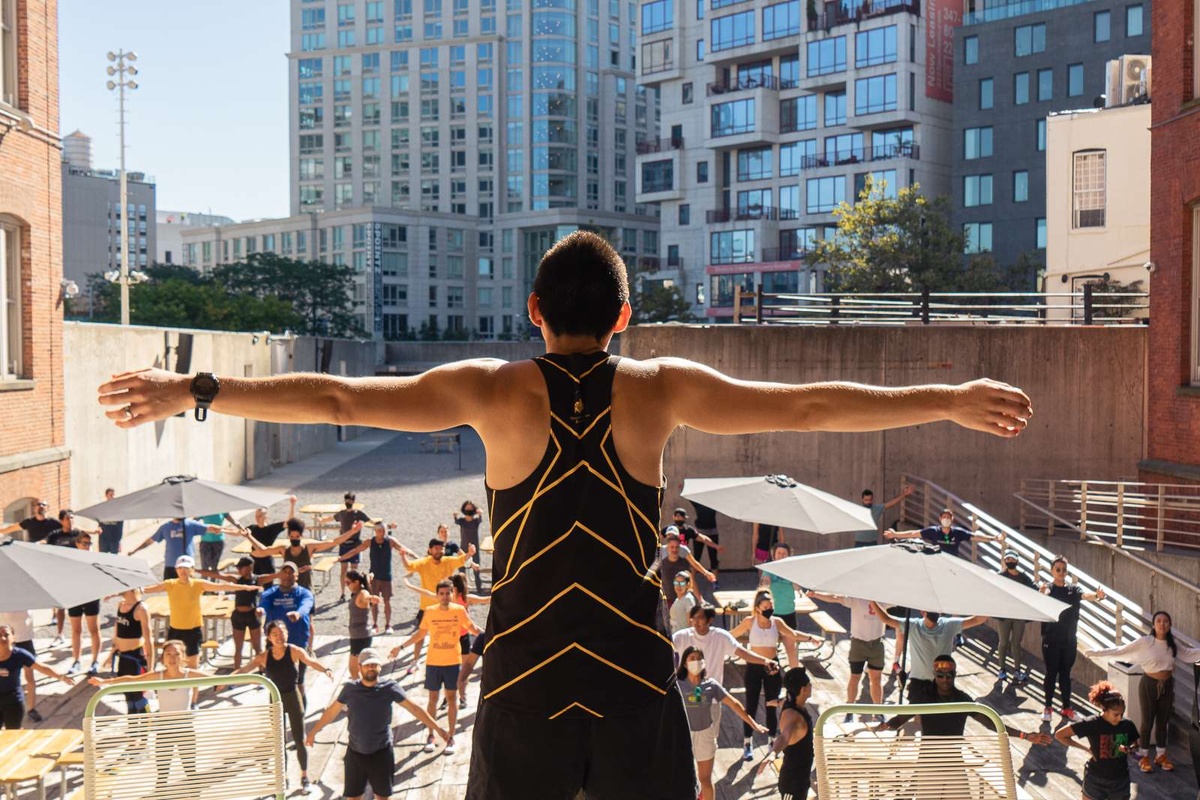 A person stands with arms spread above a courtyard where people stretch before a run.