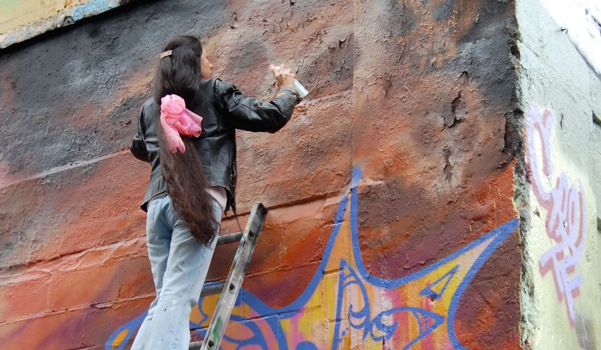 Lady pink spray paints a wall as she stands on a ladder