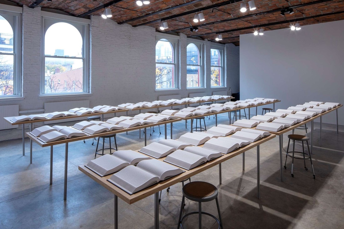 Open books with bright white pages filled with small text are laid out in rows across six long tables with stools on either side. The room has large arch way windows and a brick ceiling with bright spotlights suspended from it.