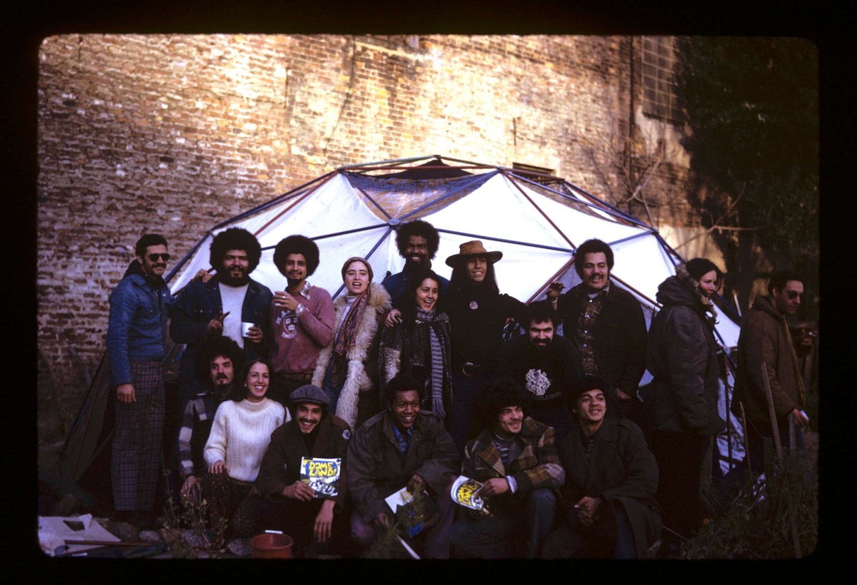 A vintage group photograph in front of a luminous geometric dome. Some are squatting in the front holding flyers that read "DOME LAND!" in bright yellow.
