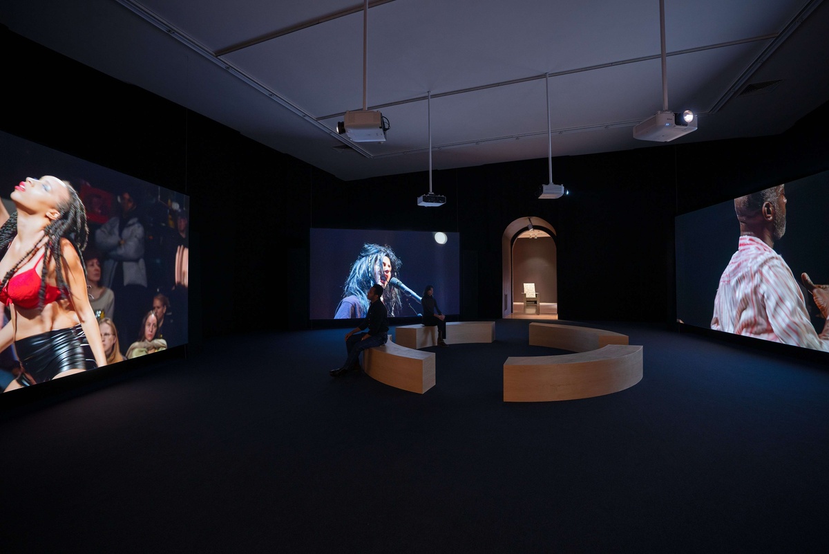 Three screens display pre-recorded dance performances in a dark room.