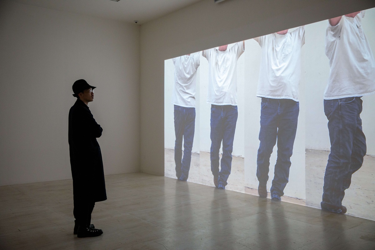 A figure in a dark outfit watches a four paneled projected video of a person wearing a white t-shirt and baggy dark blue jeans.