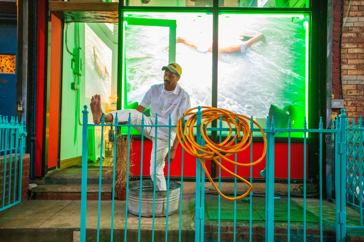 A person, dressed in all white, stands with on foot in a metal tub and the other foot on top of a blue fence.