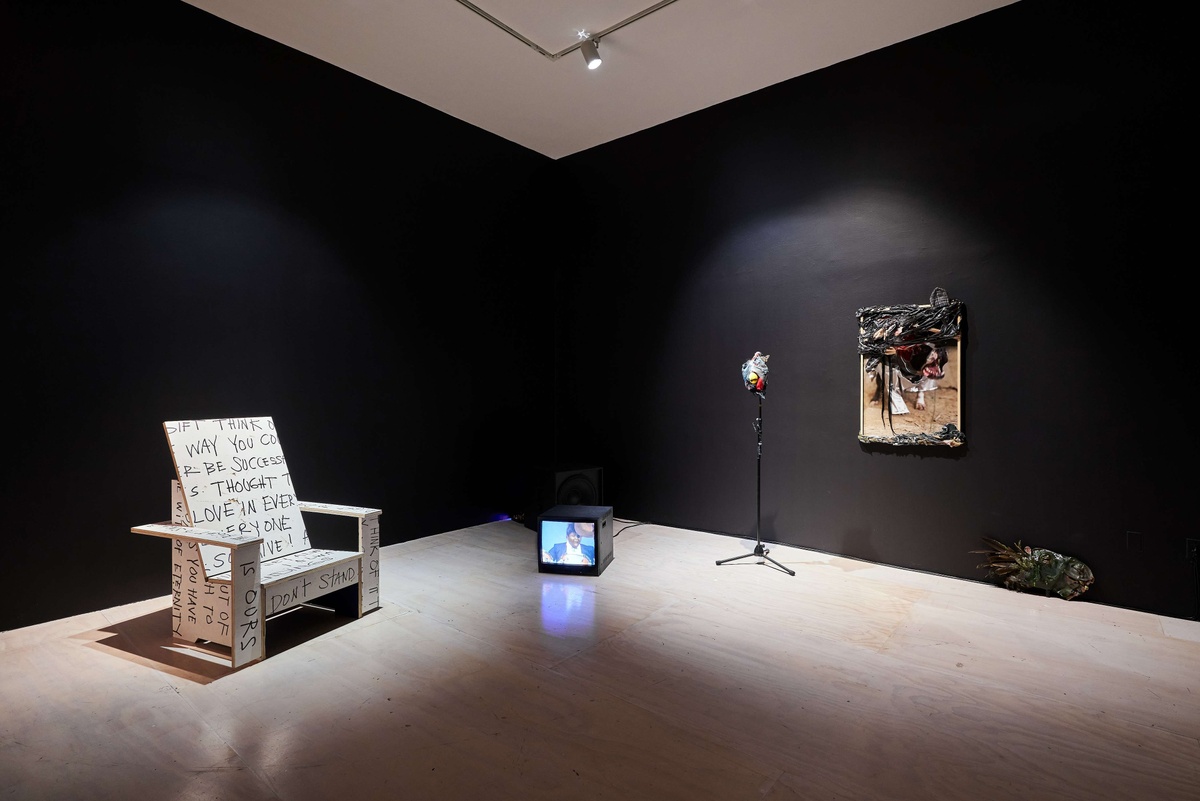 A chair, a television set, a chicken's head placed on a microphone stand, and a painting displayed in a dark room.