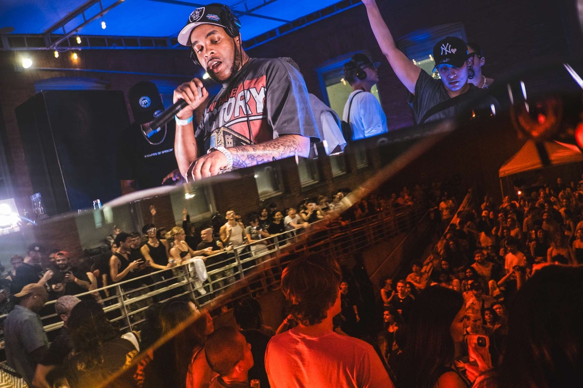 A DJ stands above a reflect booth which shows a joyful crowd at MoMA PS1's Warm Up. 