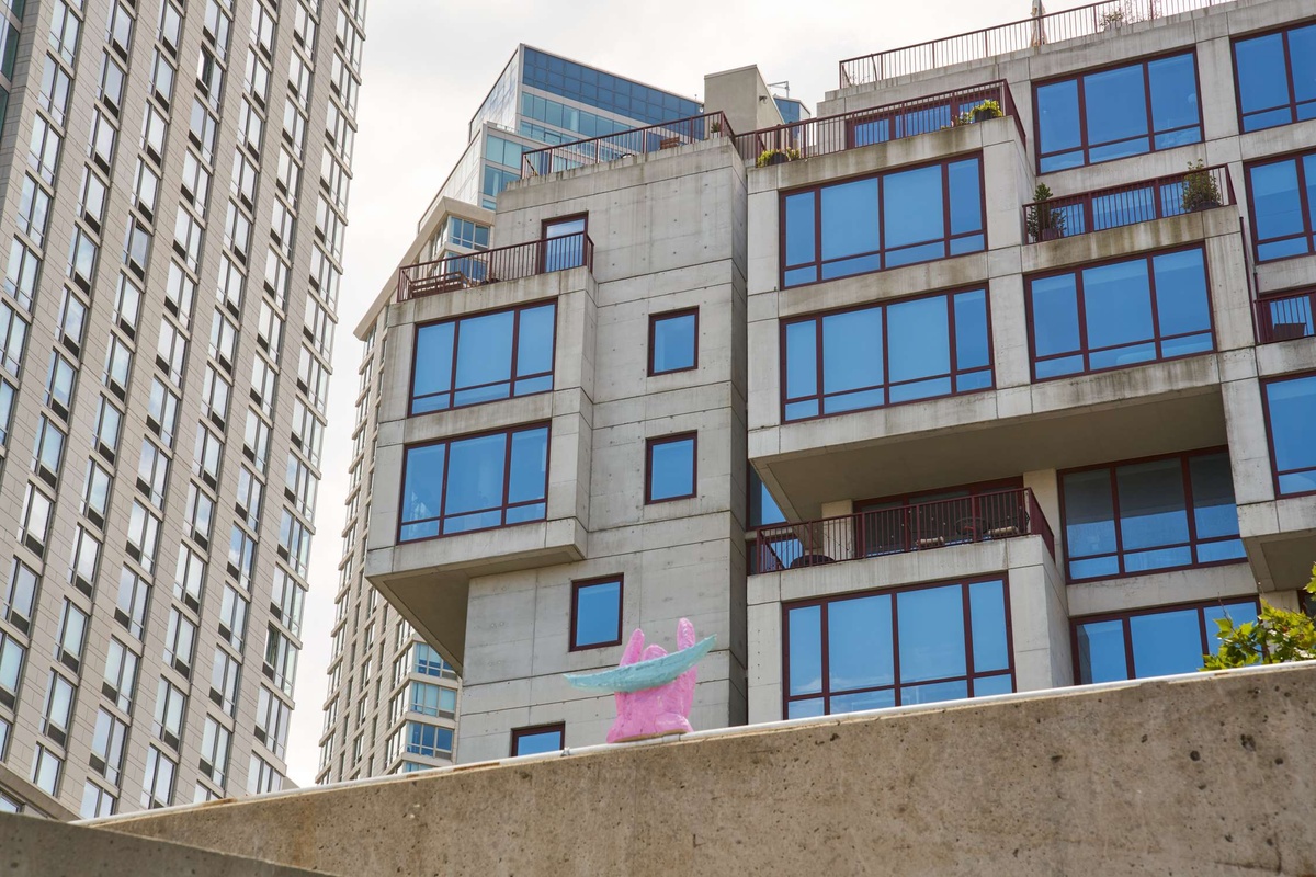 A knome-like baby pink & blue sculpture sits on a wall against large building. The sculpture has its arms raised to the sky.