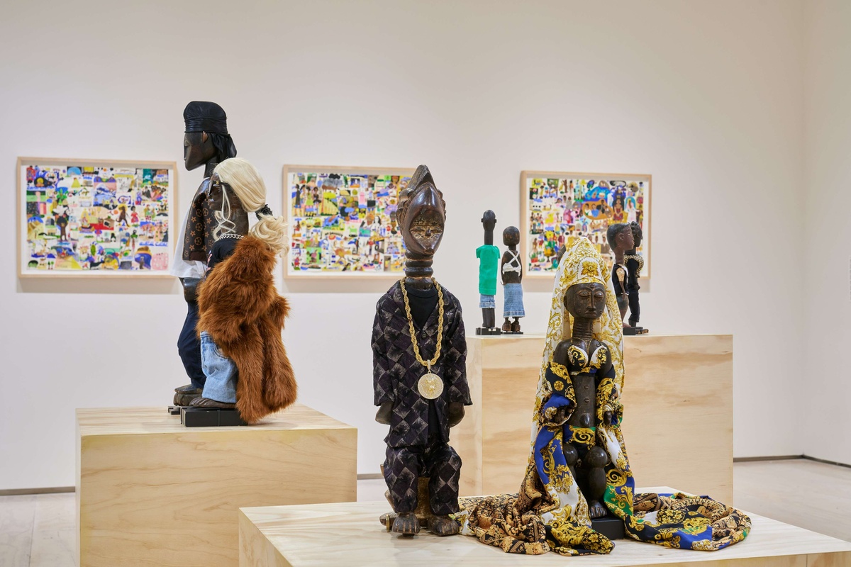 Multiple African wooden statues dressed in contemporary garb sitting on wooden pedestals, surrounded by artwork.