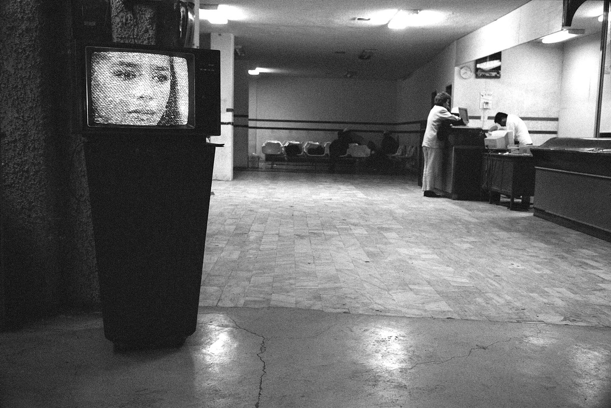 A photo by Reynaldo Rivera depicts a nearly empty bus station. In the foregound, a static-filled TV displays the tear-filled eyes of a woman.