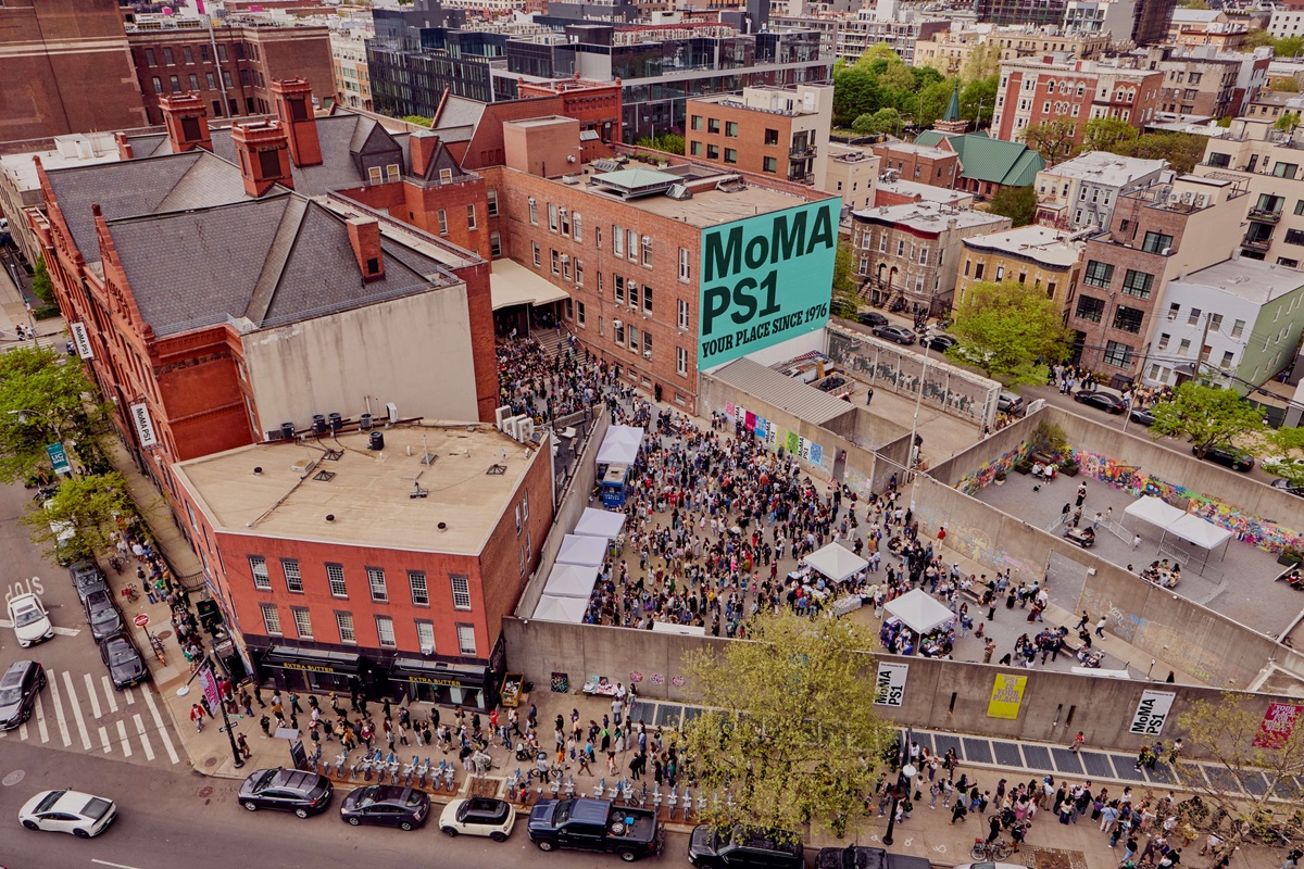 Aerial view of MoMA PS1 in Long Island City, Queens, with a large teal "MoMA PS1 / Your Place Since 1976" mural on the building's exterior wall. A dense crowd fills the courtyard and street below for a block party, with white tents, a graffiti-covered concrete wall, and a crowd lining the sidewalk.
